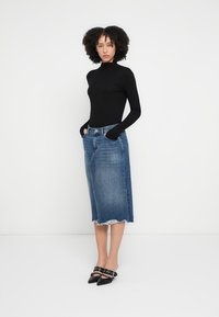 Mustad pikkade varrukatega kõrge kaelusega pluus, sinine denimmidiseelik franseeritud äärega, kantud mustade teravate kandadega kingadega, seistes valge tausta ees.