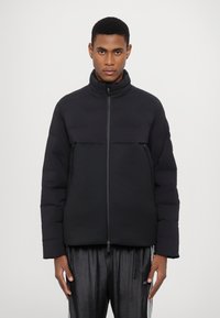 Puffer jacket in black, with a high collar and a zip closure. Features a two-tone design with a smooth upper and textured lower section.