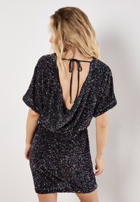 Schwarzes Paillettenkleid mit tiefem V-Rücken, kurzen Ärmeln und gerafftem Taillenschnitt. Mit schwarzer Schleifenverschluss am Rücken. Funkelnde Textur über das gesamte Kleid.