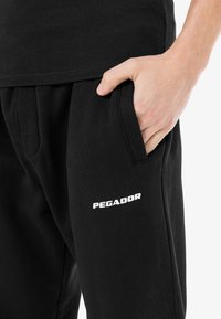 Pantaloni de trening negri cu o textură moale, dotati cu un buzunar lateral și un logo alb "PEGADOR" pe piciorul stâng. Talie elastică vizibilă.