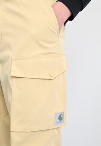Beige cargo bukser laget av slitesterk stoff, med en sidelomme med klaff og trykknapp, samt et Carhartt-logo-patch.