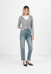 Jonge vrouw staat met haar hand in de zak, gekleed in een grijze zip-up hoodie, een witte grafische T-shirt, blauwe jeans en zwarte puntige hakken.