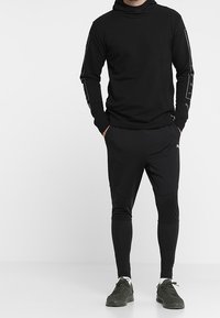 Top negro de manga larga con cuello alto, que presenta un tejido texturizado y detalles de logo en las mangas, combinado con pantalones negros ajustados y zapatillas deportivas.