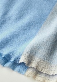 Tessuto in misto cashmere blu chiaro con tonalità variabili, caratterizzato da una consistenza morbida e bordi frangiati, esposto in un arrangiamento piegato.