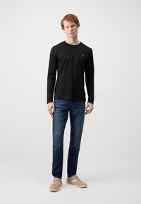 Chemise noire à manches longues avec un col rond, assortie à un jean en denim bleu et des baskets beiges avec des accents blancs.