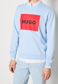 Sweat-shirt bleu clair avec un graphique rectangulaire rouge affichant "HUGO" en lettres noires épaisses. Comprend des poignets et un ourlet côtelés.