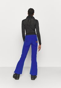 Niebieskie spodnie narciarskie z rozszerzanymi nogawkami, wzmocnionym szwem i zamykanymi na zamek kieszeniami, zestawione z czarną, wzorzystą bluzką z długim rękawem. Widoczne czarne buty.