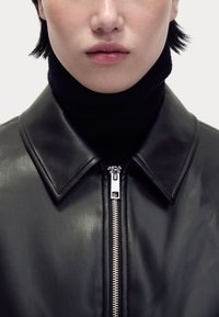 Schwarze Lederjacke mit klassischem Kragen und silbernem Reißverschluss. Über einem schwarzen Rollkragenpullover getragen, der eine glatte Textur zeigt.