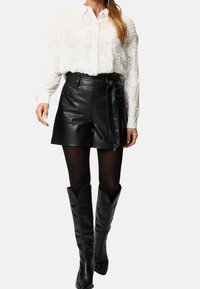 Chemise blanche avec des embellissements floraux texturés, shorts taille haute en cuir noir avec poches latérales et une ceinture nouée, bottes noires hauteur genou.