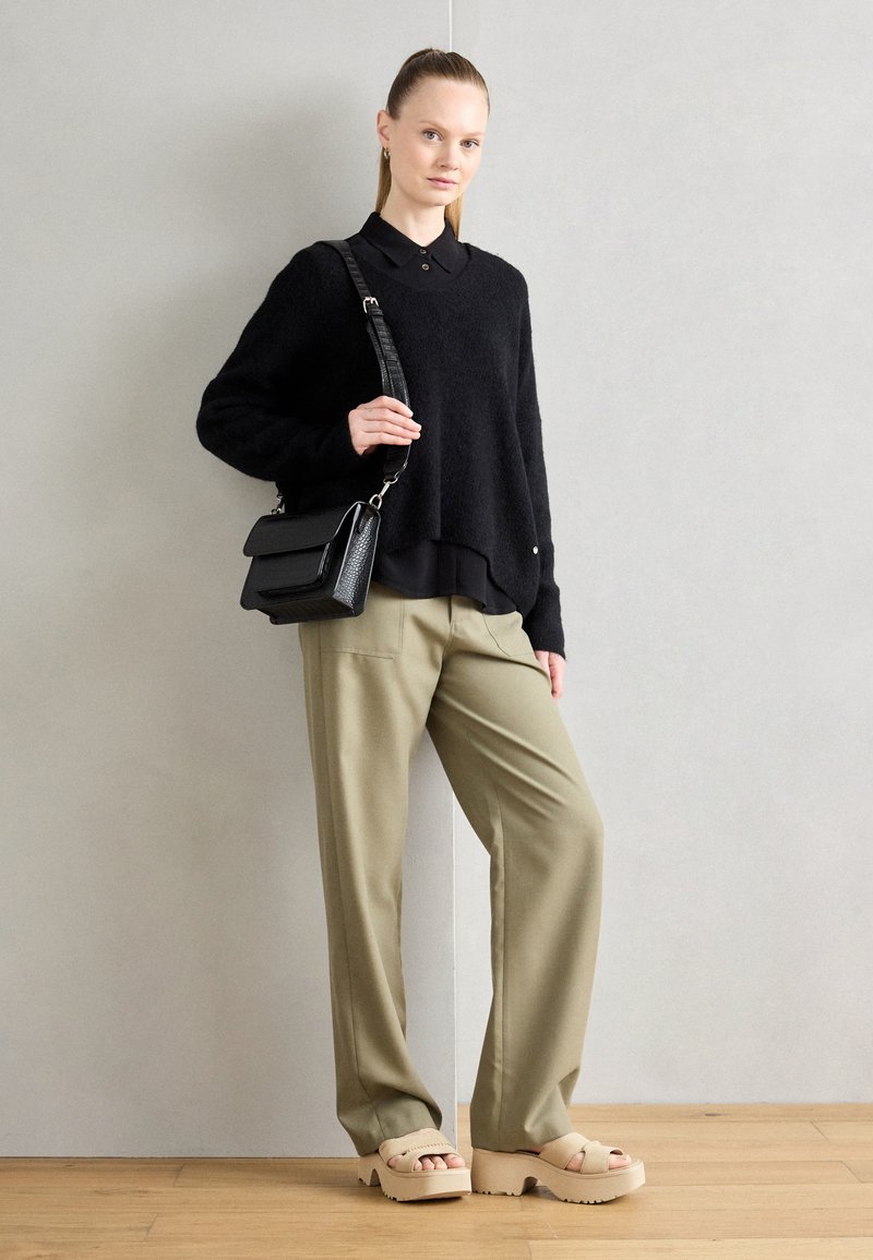 Schwarzer, kuscheliger Pullover über einem schwarzen Hemd mit Kragen, olivgrünen weiten Hosen und beigen Plateausandalen. Schwarze Umhängetasche mit strukturierten Details.
