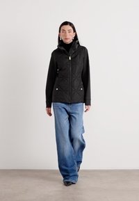 Een vrouw loopt zelfverzekerd, gekleed in een zwart gewatteerd vest over een zwart top en losse blauwe jeans, tegen een minimalistische achtergrond.