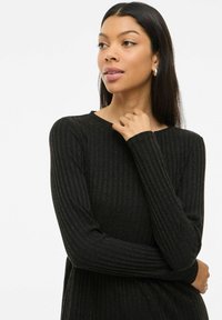 Femme avec de longs cheveux noirs portant un haut à manches longues noir à côtes, regardant légèrement sur le côté avec une main près de son cou.