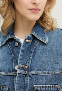 Kurtka jeansowa w jasnoniebieskim kolorze, z srebrnymi guzikami i kontrastującym szwem. Posiada kołnierz z klapą oraz przednie kieszenie.