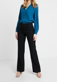 Bluzka w kolorze morskim z dekoltem w serek i długimi rękawami, zestawiona z czarnymi spodniami z wysokim stanem, ozdobionymi guzikami i rozszerzanymi nogawkami.