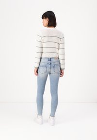 Fehér és zöld csíkos, kötött pulóver, világoskék, skinny farmernadrággal párosítva. A modell fehér sportcipőt visel, hátulról készült nézet.