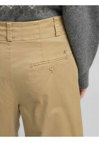 Pantaloni beige con una texture liscia, caratterizzati da una tasca posteriore con chiusura a bottone e un dettaglio logo sottile. La vita è piatta con passanti per cintura.