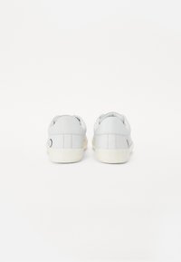 Pár bílých nízkých tenisek zobrazených zezadu s minimálním šitím a světlými podrážkami na bílém pozadí.