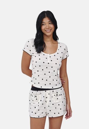 Donna che indossa una maglietta bianca a maniche corte e pantaloncini con pois neri e piccole tasche frontali, sorridente e che guarda di lato.