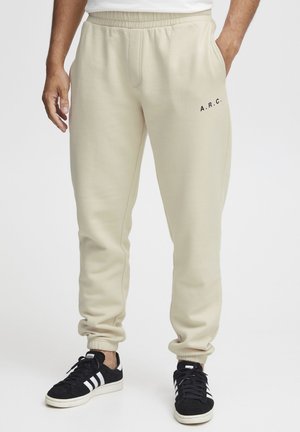 Beige bomulls joggebukser med elastisk linning og ribbede mansjetter. Har "A.R.C." i svart på venstre lår. Brukt med svarte joggesko.
