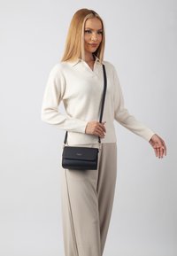 Borsa a tracolla nera in pelle con superficie texturizzata, hardware dorato e chiusura a patta. Il modello indossa un maglione color crema e pantaloni beige.