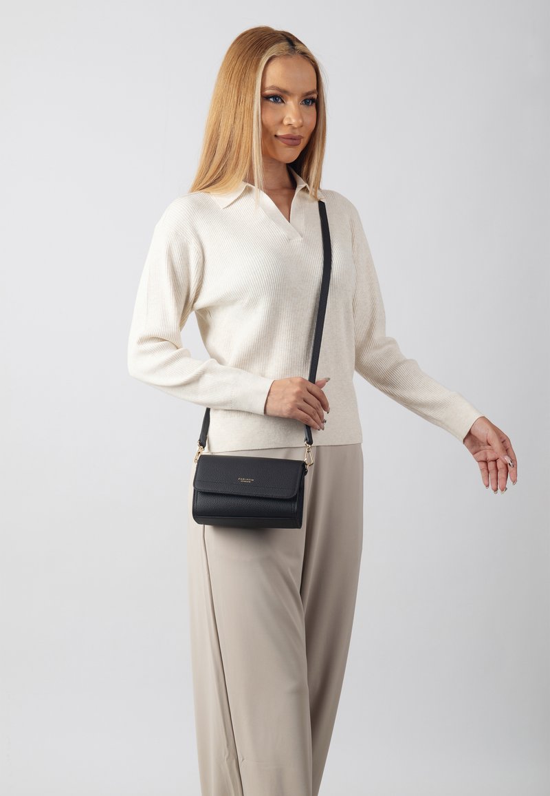 Borsa a tracolla nera in pelle con superficie texturizzata, hardware dorato e chiusura a patta. Il modello indossa un maglione color crema e pantaloni beige.