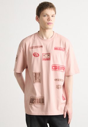 T-shirt surdimensionné rose clair en coton, ornée de divers patchs graphiques rouges et bruns avec des noms de pays et des logos.