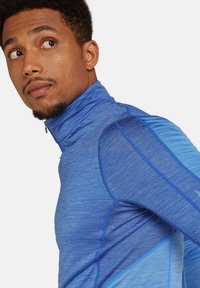 Camiseta atlética de mangas largas y cuello alto en tonos variados de azul, con tejido texturizado, cremallera y detalles de panel contrastantes en las mangas.