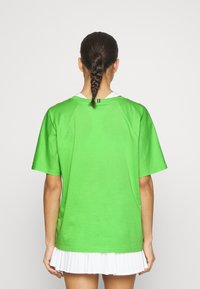 Camiseta verde de algodón con mangas cortas, corte relajado y cuello acanalado. Camiseta blanca visible por debajo y falda blanca con pliegues en la parte inferior.