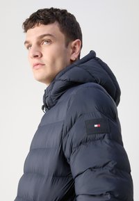 Mornarska puf jakna ima kapuljaču, kviltanu teksturu i gumeni logo na rukavu. Materijal je gladak i lagan.