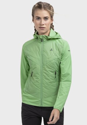 Frau mit geflochtenem Haar, die eine hellgrüne Kapuzenjacke und schwarze Hose trägt, steht vor einem schlichten, hellen Hintergrund.