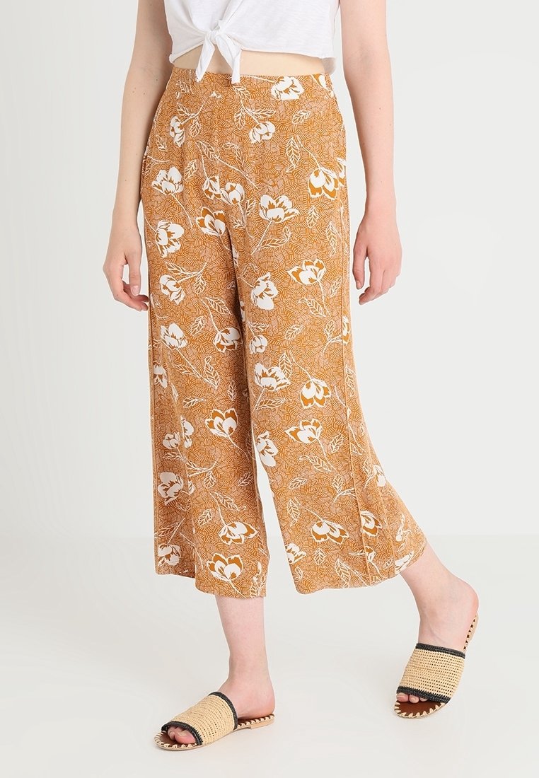 Pantalons culotte en tissu marron doré avec un motif floral blanc. Coupe ample avec une taille mi-haute. Portés avec des sandales ouvertes à bout plat.