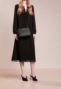 Dlouhé černé šaty s volánkovými detaily, lemem s bambulkami a dlouhými rukávy. Kombinovány s černou kabelkou a zdobenými lodičkami na vysokém podpatku.