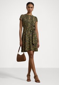 Abito floreale verde con maniche corte, scollo plissettato, cintura in vita e orlo sopra il ginocchio. Abbinato a una borsa marrone e sandali.