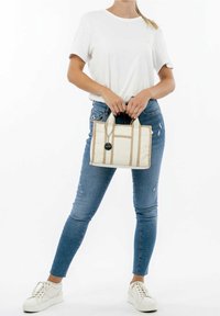 Borsa a tracolla in tela beige con strisce marroni, dotata di un'etichetta rotonda. Indossata con jeans blu e sneakers bianche, abbinata a una t-shirt bianca semplice.