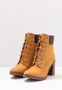 Botas de tobillo de cuero color tostado con un acento marrón en el cuello, cordones amarillos y un tacón grueso. Suela de goma texturizada con patrón de tracción.