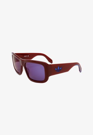 Occhiali da sole rettangolari con lenti viola, montatura spessa rossa e logo Adidas blu sulla stanghetta.
