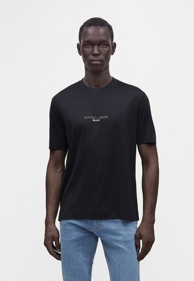 Мъж, облечен в черна тениска Emporio Armani Milano и светлосини дънки, стои пред обикновен светъл фон.