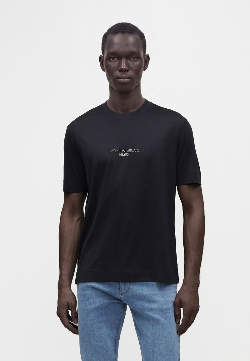 Άνδρας που φοράει μαύρο T-shirt Emporio Armani Milano και ανοιχτό μπλε τζιν, στέκεται μπροστά σε απλό φωτεινό φόντο.