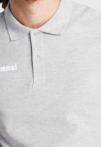 Polo gris en coton, avec un col, une patte à deux boutons et un logo blanc sur la poitrine. Manches courtes visibles.