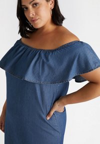 Denimkleid mit schulterfreiem Schnitt, rüschiertem Ausschnitt, kurzen Ärmeln und seitlichen Taschen. Dunkelblaues Gewebe mit glatter Textur.