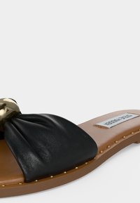 Μαύρο δερμάτινο σανδάλι τύπου slip-on με διακόσμηση από χρυσά καρφιά και μια μεγάλη χρυσή αγκράφα στο μπροστινό λουράκι, με την ετικέτα "Steve Madden" στην εσωτερική σόλα.