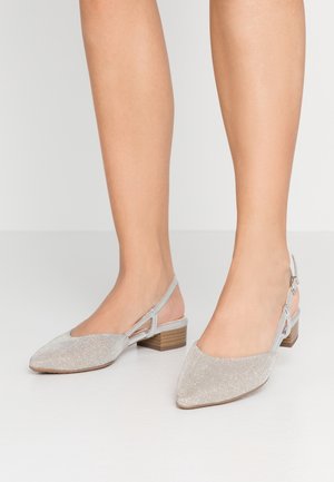 Femme portant des chaussures argentées à bout pointu, à bride arrière, avec des talons blocs bas et des brides à la cheville, debout sur une surface blanche lisse.