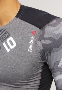 Šedé a černé sportovní tričko s krátkými rukávy, s logem Reebok, maskáčovým vzorem a síťovou texturou na ramenech.