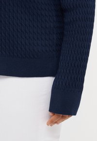 Marineblauw gebreid sweater met een gestructureerd kabelpatroon, ribbels aan de mouwen en de onderkant. Draag het over witte broek, met een close-up van de mouw.