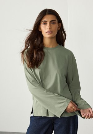 Kobieta o długich ciemnych włosach w zielonej bluzce z długim rękawem i ciemnych dżinsach, poprawiająca rękaw na jednolitym jasnym tle.