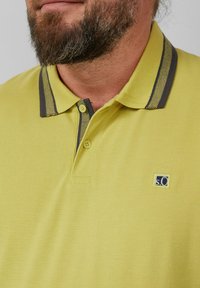 Gelbes Poloshirt aus strukturierter Baumwolle, mit kontrastierendem grauen Kragen und zwei Knöpfen. Es hat ein kleines blaues Logo auf der Brust.