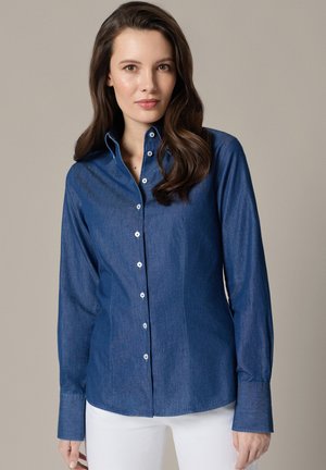 Kobieta z długimi ciemnymi włosami, ubrana w zapinaną na guziki niebieską jeansową koszulę i białe spodnie, stoi na tle jednolitego szarego tła.