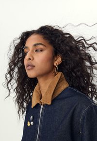 Veste en denim avec un col en velours côtelé beige, fermeture éclair argentée, et trois pin's sur la poitrine. Le modèle a des cheveux bouclés et fait légèrement face à gauche.
