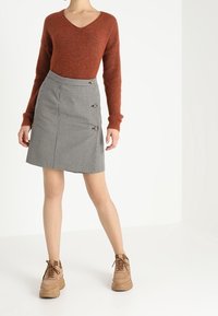 Pull tricoté de couleur rouille avec col en V ; jupe à carreaux gris avec trois boutons latéraux ; chaussures à lacets couleur beige. Fond uni.