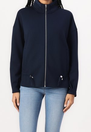Personne portant une veste zippée bleu marine à col montant et un jean bleu clair sur un fond blanc uni.